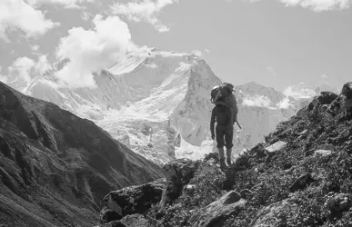 Een persoon met een grote rugzak staat op een rotsachtig bergpad en kijkt in de richting van een met sneeuw bedekte bergtop in de verte, onder een gedeeltelijk bewolkte hemel.
